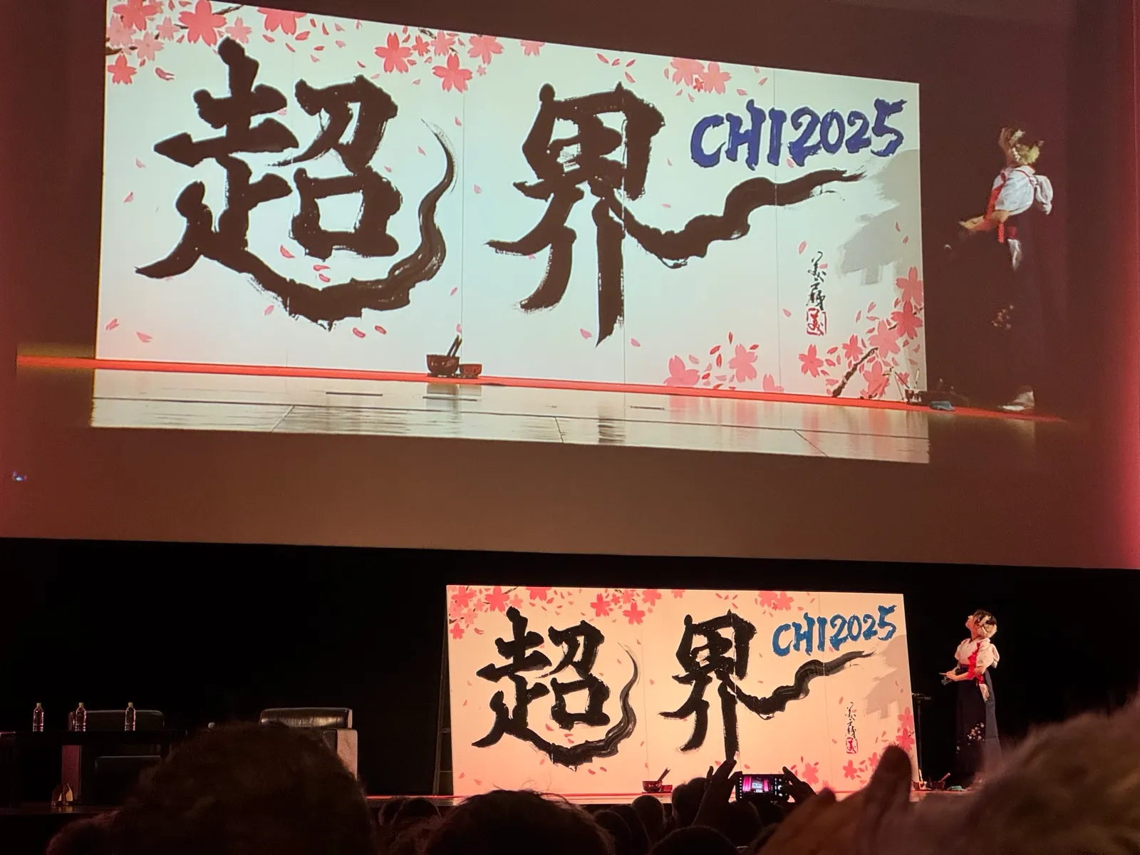 A calligrapher on stage beside a large brush-lettered banner reading CHI2025 with cherry blossoms, also shown on a big screen above.