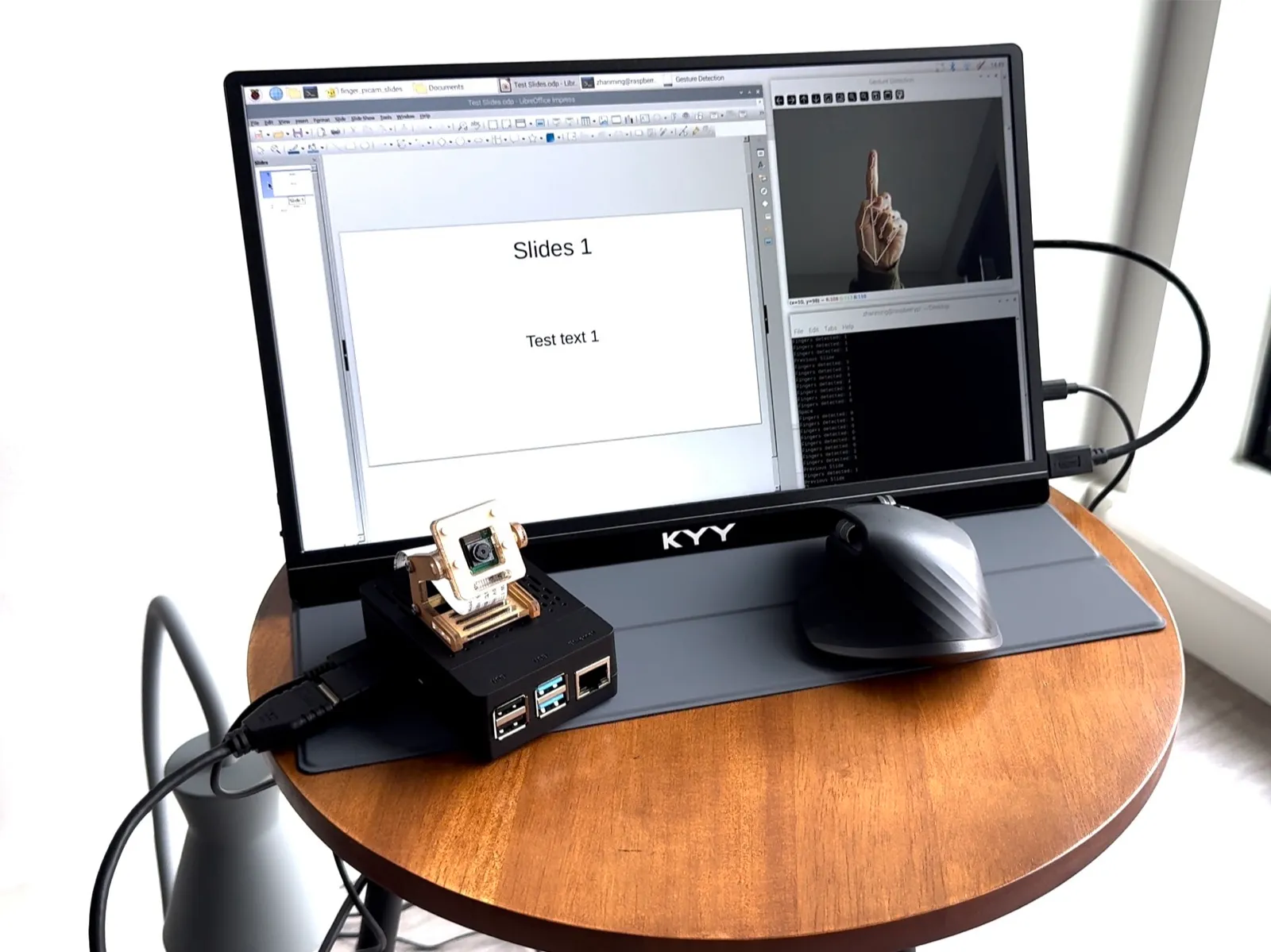 The prototype on a side table: a Raspberry Pi and camera beside a monitor showing slides and the gesture-detection window